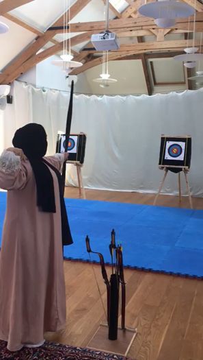 A woman in a hijab, seen from behind, aiming a bow towards targets in an indoor hall.