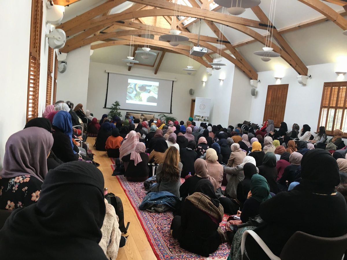 Women in hijab seated in a packed hall, mostly seen from behind, facing a projector screen.