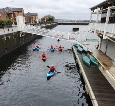 Kayakers far off on calm water, with a white footbridge and a wooden dock beside them.